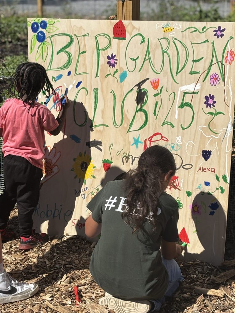 Two youth paint a wooden BEP Garden Club sign outdoors, faces turned away.