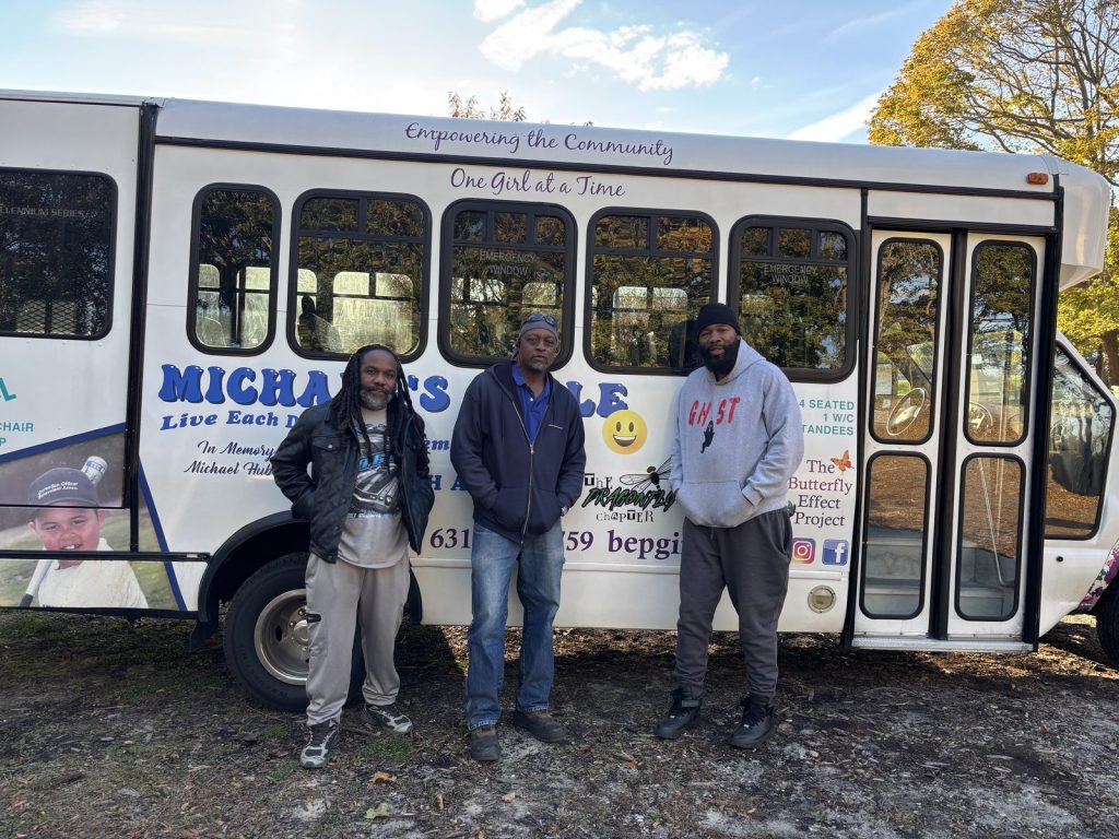 Three men stand in front of a white Butterfly Effect Project branded shuttle bus.