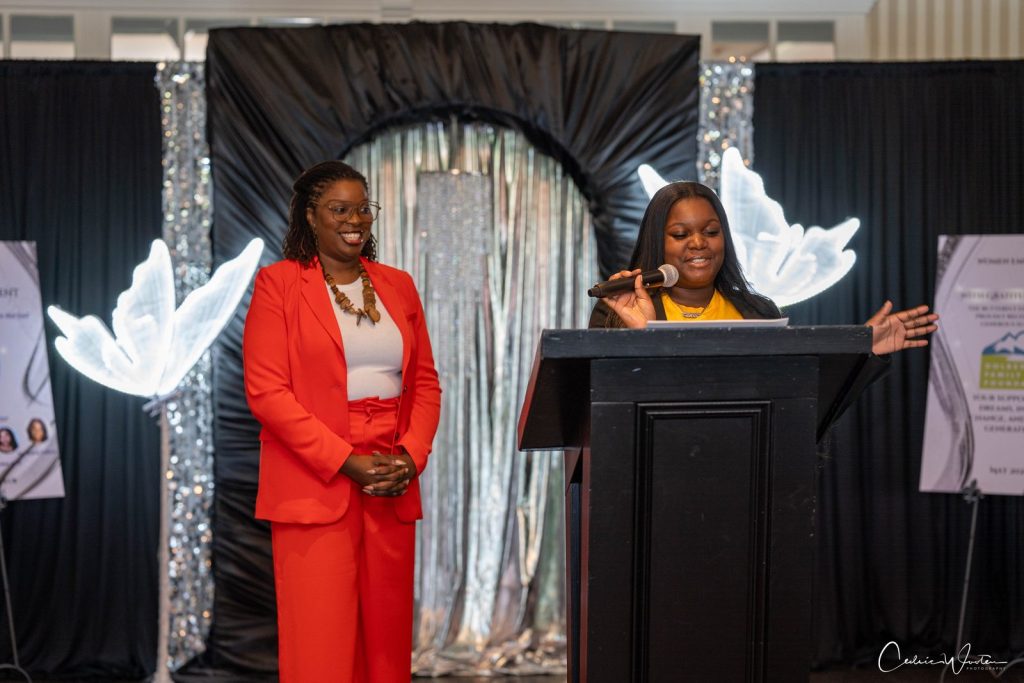 Ms. Tia in a red suit smiles beside a young speaker at a podium framed by glowing white butterfly wing props.