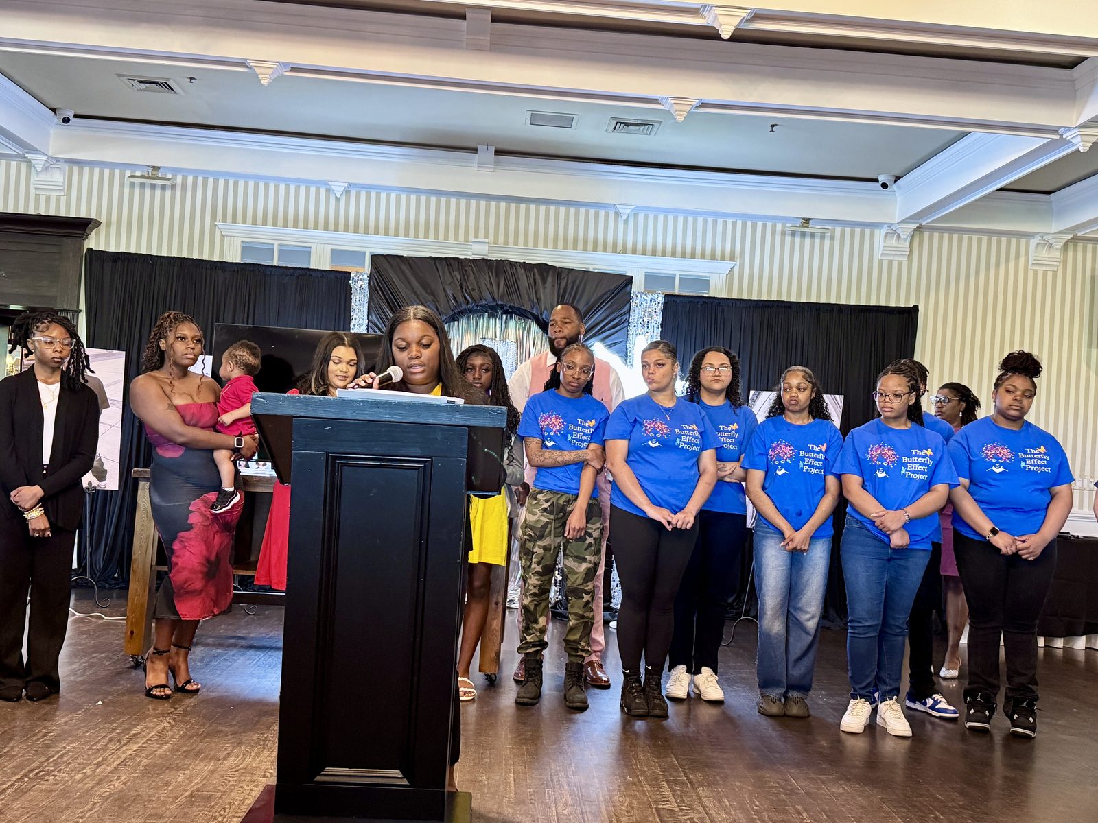 A speaker stands at a podium while youth in matching blue Butterfly Effect shirts stand behind her.