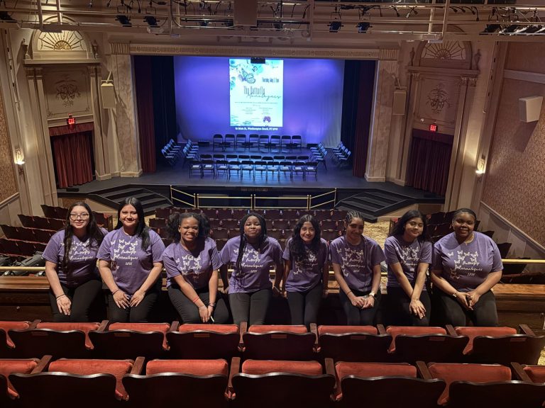 Girls in purple Butterfly Effect shirts sit on a stage edge in a large auditorium.