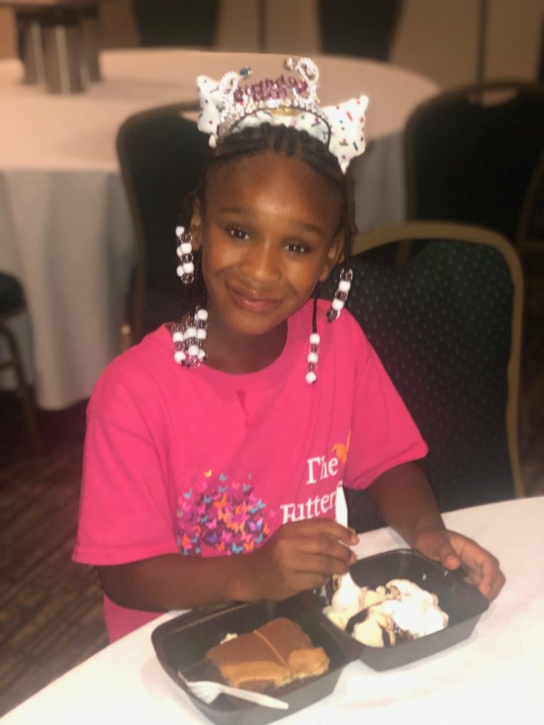 A girl in a pink shirt and tiara sits smiling at a banquet table.