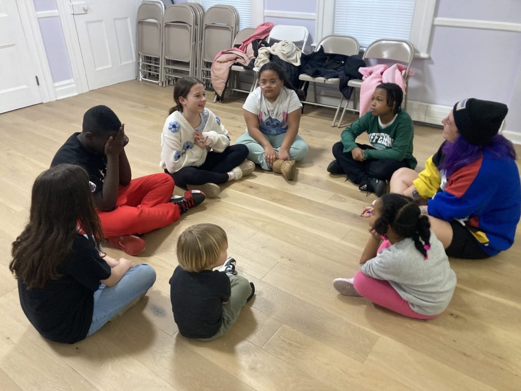 Children and a mentor with purple hair sit cross-legged in a circle on a wooden floor.