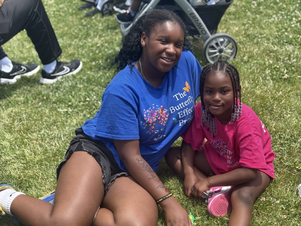 An older girl in a blue project shirt sits on the grass smiling with a younger girl in pink.