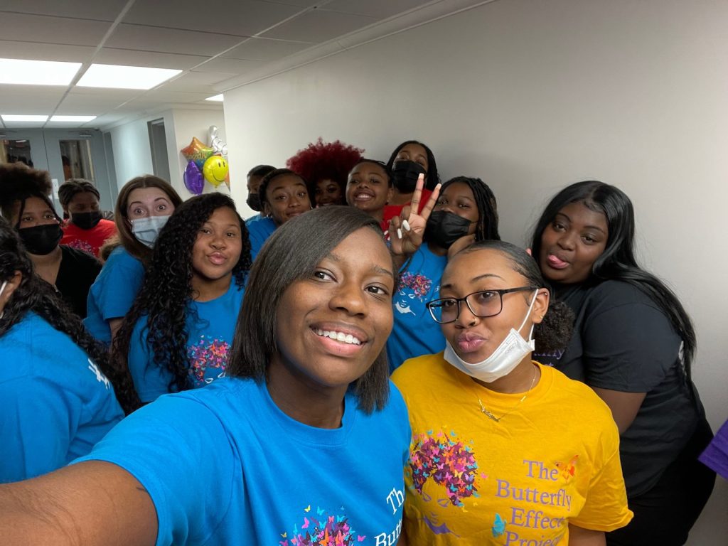 Young women smile up into a group selfie; one wears a bright blue Butterfly Effect shirt.