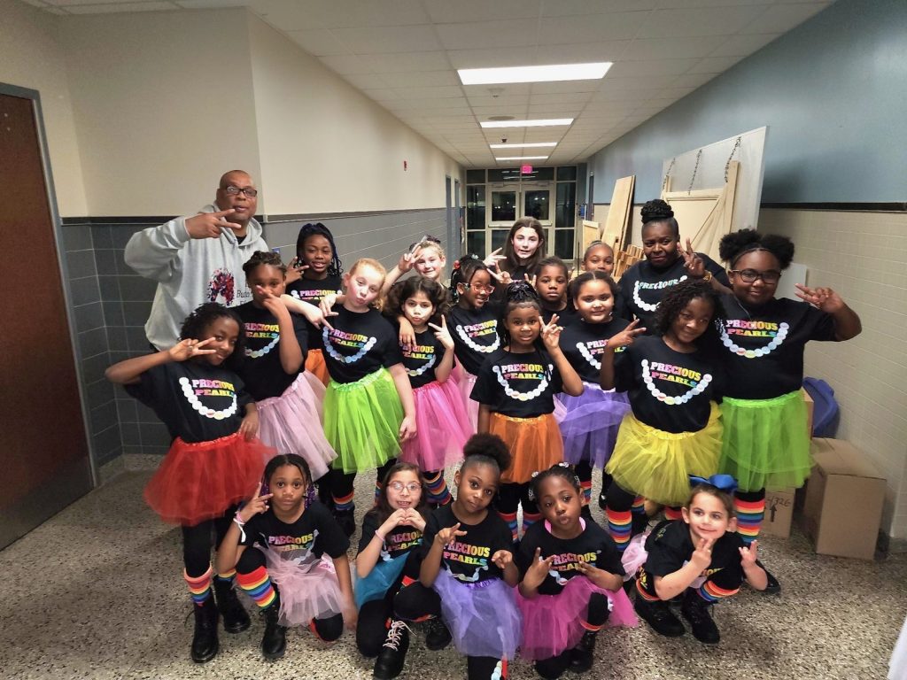A large group of girls in black project shirts and colorful tutus pose together in a hallway.