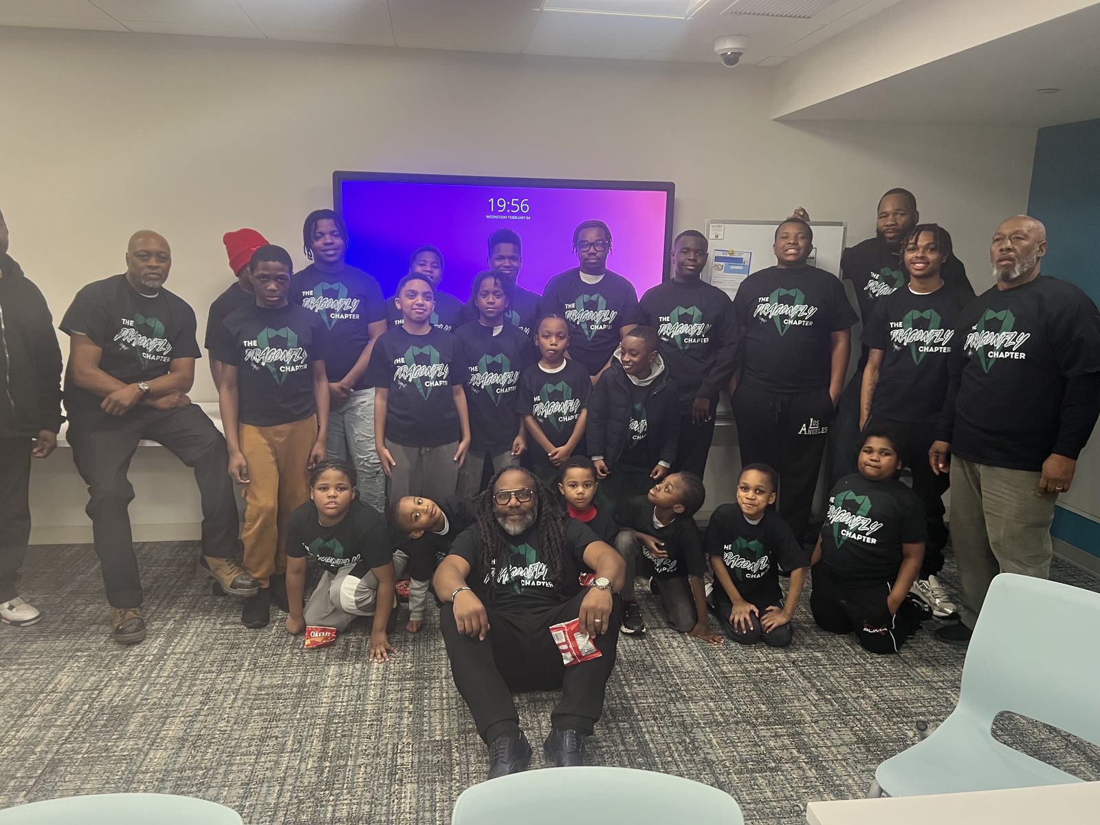 A large indoor group of children and adults in matching black logo shirts poses in rows.