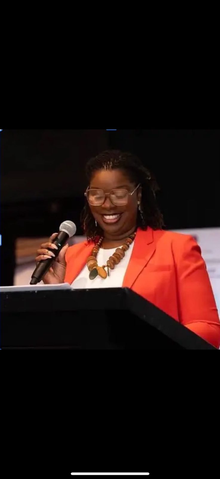 Ms. Tia smiles while speaking into a microphone at a podium, wearing an orange blazer and a wooden bead necklace.